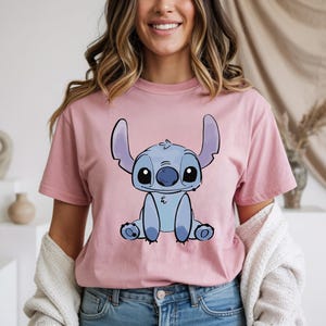 Puede incluir: Una camiseta rosa claro con una ilustraci&oacute;n de dibujos animados de Stitch de Lilo & Stitch. El personaje azul est&aacute; sentado con orejas grandes y ojos grandes. La camiseta es de color s&oacute;lido y tiene mangas cortas.