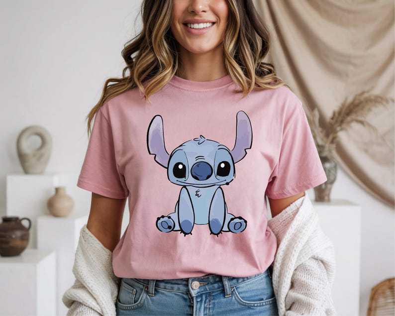 Puede incluir: Una camiseta rosa claro con una ilustraci&oacute;n de dibujos animados de Stitch de Lilo & Stitch. El personaje azul est&aacute; sentado con orejas grandes y ojos grandes. La camiseta es de color s&oacute;lido y tiene mangas cortas.