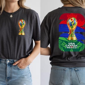 May include: Dark gray t-shirt featuring a gold trophy with colorful accents on the front and back. The back design includes the text "USA CANADA MEXICO" and the number "26". The shirt is made of a soft material.