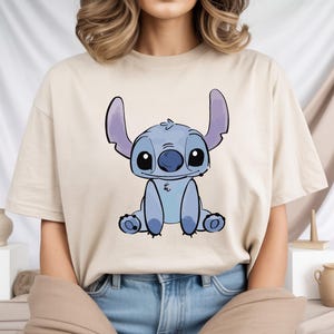 Puede incluir: Camiseta beige con una ilustraci&oacute;n de dibujos animados de Stitch, un personaje alien&iacute;gena azul con orejas grandes y una expresi&oacute;n amigable. El gr&aacute;fico est&aacute; centrado en la parte delantera de la camiseta. La camiseta est&aacute; hecha de una tela suave y clara.