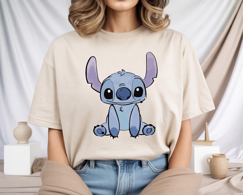 Puede incluir: Camiseta beige con una ilustraci&oacute;n de dibujos animados de Stitch, un personaje alien&iacute;gena azul con orejas grandes y una expresi&oacute;n amigable. El gr&aacute;fico est&aacute; centrado en la parte delantera de la camiseta. La camiseta est&aacute; hecha de una tela suave y clara.
