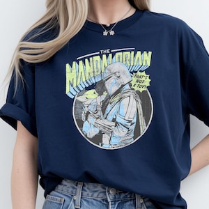 May include: Navy blue t-shirt featuring a graphic of The Mandalorian holding Grogu, with the text "THAT'S NOT A TOY!". The design is in a comic book style with blue, yellow, and white accents. The shirt has rolled-up sleeves.