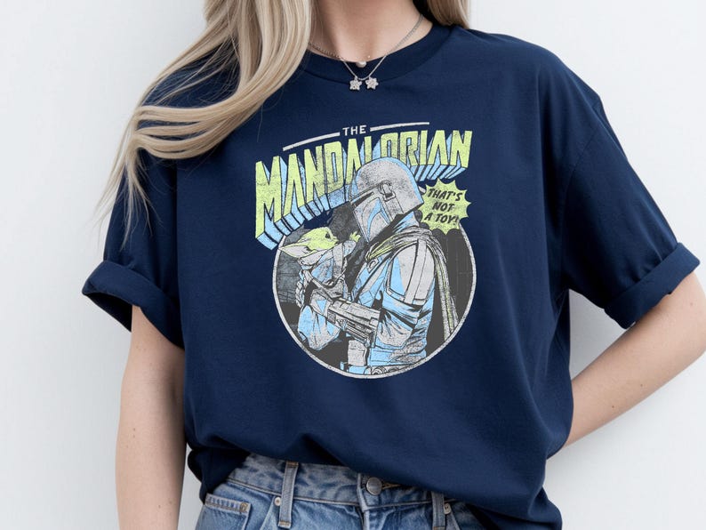 May include: Navy blue t-shirt featuring a graphic of The Mandalorian holding Grogu, with the text "THAT'S NOT A TOY!". The design is in a comic book style with blue, yellow, and white accents. The shirt has rolled-up sleeves.