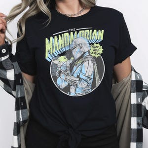 May include: Black t-shirt featuring a graphic of The Mandalorian holding Grogu, with the text "THE MANDALORIAN" in green and blue, and "THAT'S NOT A TOY!" in yellow. The shirt is worn with a black and white plaid shirt.