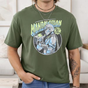 May include: Olive green t-shirt featuring a graphic from The Mandalorian. The design includes the text "THE MANDALORIAN" and "THAT'S NOT A TOY!" with an illustration of the Mandalorian and Grogu. The shirt is a crew neck and has short sleeves.