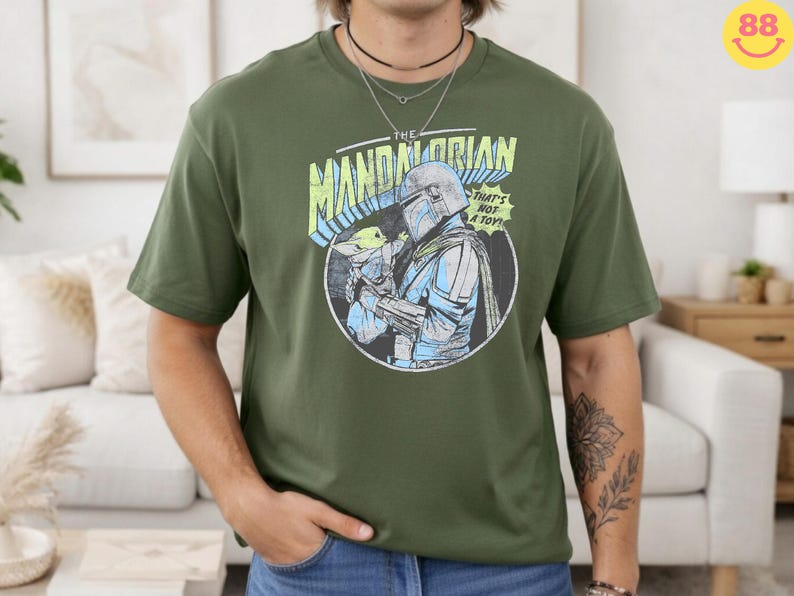May include: Olive green t-shirt featuring a graphic from The Mandalorian. The design includes the text "THE MANDALORIAN" and "THAT'S NOT A TOY!" with an illustration of the Mandalorian and Grogu. The shirt is a crew neck and has short sleeves.