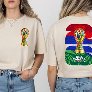 May include: Beige t-shirt featuring a colorful trophy graphic on the front and back. The front has a small trophy, while the back displays a larger trophy design with the text "USA CANADA MEXICO". The shirt is made of a soft material.