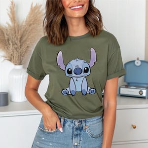 Puede incluir: Camiseta verde oliva con una ilustraci&oacute;n de dibujos animados de Stitch de Lilo & Stitch. El personaje est&aacute; representado en tonos azules y blancos, con ojos grandes y orejas prominentes. La camiseta tiene mangas cortas y un corte holgado.