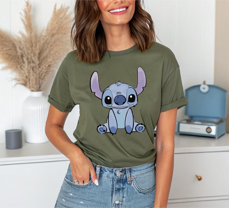 Puede incluir: Camiseta verde oliva con una ilustraci&oacute;n de dibujos animados de Stitch de Lilo & Stitch. El personaje est&aacute; representado en tonos azules y blancos, con ojos grandes y orejas prominentes. La camiseta tiene mangas cortas y un corte holgado.