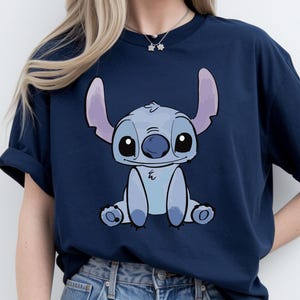 Puede incluir: Camiseta azul marino con una ilustraci&oacute;n de dibujos animados de Stitch, una criatura extraterrestre azul con orejas grandes y ojos grandes. El gr&aacute;fico est&aacute; centrado en la parte delantera de la camiseta. La camiseta es de material suave.