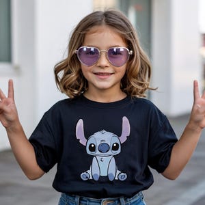 Puede incluir: Una ni&ntilde;a lleva gafas de sol moradas en forma de coraz&oacute;n y una camiseta negra con un personaje de dibujos animados. La ni&ntilde;a sonr&iacute;e y hace el signo de la paz con ambas manos. La camiseta tiene un dise&ntilde;o azul y morado.