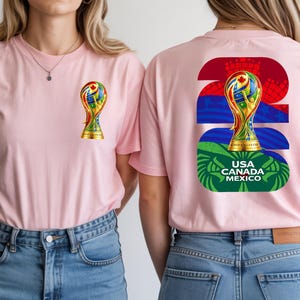 May include: Two pink t-shirts with a colorful graphic of a trophy on the front and back. The back of the shirt has the number 26, a trophy graphic, and the text "USA CANADA MEXICO". The shirts are worn with blue jeans.