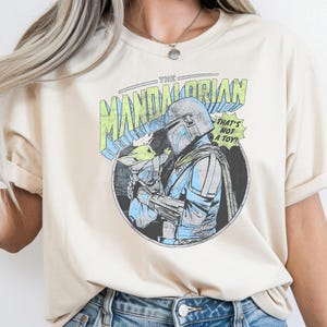 May include: Cream-colored t-shirt featuring a graphic of The Mandalorian holding Grogu, with text reading "THE MANDALORIAN" and "THAT'S NOT A TOY!". The design is in a comic book style with blue, green, and black accents. The shirt has rolled-up sleeves.