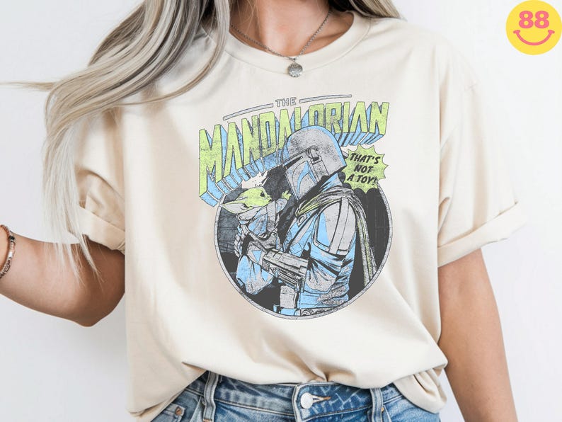 May include: Cream-colored t-shirt featuring a graphic of The Mandalorian holding Grogu, with text reading "THE MANDALORIAN" and "THAT'S NOT A TOY!". The design is in a comic book style with blue, green, and black accents. The shirt has rolled-up sleeves.