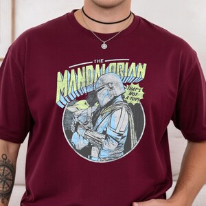 May include: Burgundy t-shirt featuring a graphic from "The Mandalorian." The design includes the text "The Mandalorian" in a yellow and blue font, along with an image of the Mandalorian and Grogu. The phrase "That's not a toy!" is also included.