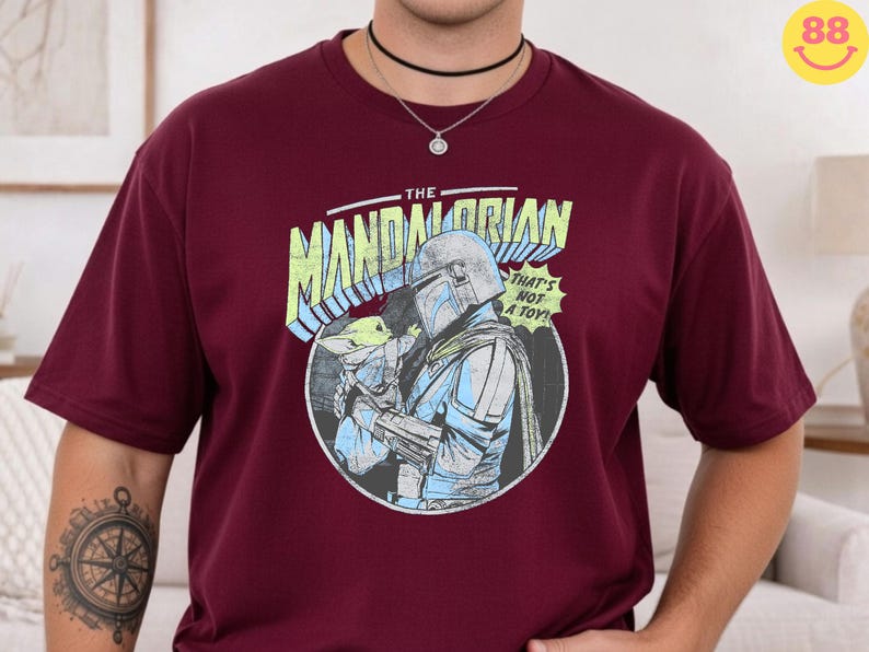 May include: Burgundy t-shirt featuring a graphic from "The Mandalorian." The design includes the text "The Mandalorian" in a yellow and blue font, along with an image of the Mandalorian and Grogu. The phrase "That's not a toy!" is also included.
