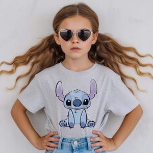 Puede incluir: Una camiseta gris claro con una imagen de dibujos animados de Stitch de Lilo & Stitch. El personaje es azul con orejas grandes y una expresi&oacute;n amigable. La persona de la imagen lleva gafas de sol en forma de coraz&oacute;n y vaqueros azules.