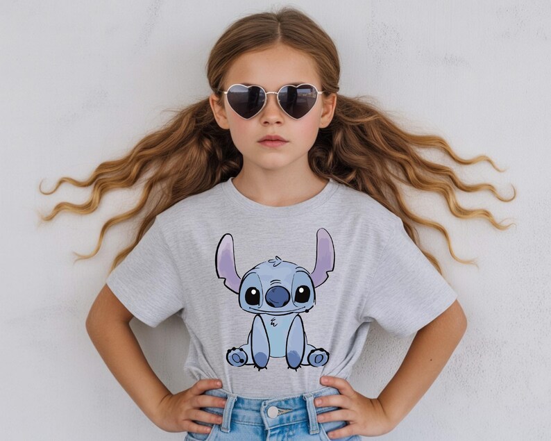 Puede incluir: Una camiseta gris claro con una imagen de dibujos animados de Stitch de Lilo & Stitch. El personaje es azul con orejas grandes y una expresi&oacute;n amigable. La persona de la imagen lleva gafas de sol en forma de coraz&oacute;n y vaqueros azules.