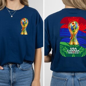May include: Navy blue t-shirt featuring a colorful trophy design on the front and back. The front displays a small trophy, while the back showcases a larger trophy with the text "USA CANADA MEXICO". The design incorporates flags and the number "26".