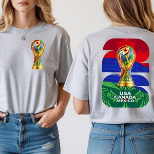 May include: A light gray t-shirt featuring a gold trophy design on the front and back. The back design includes the text "2026" and "USA CANADA MEXICO" in green, blue, and red. The trophy design incorporates various flag colors.