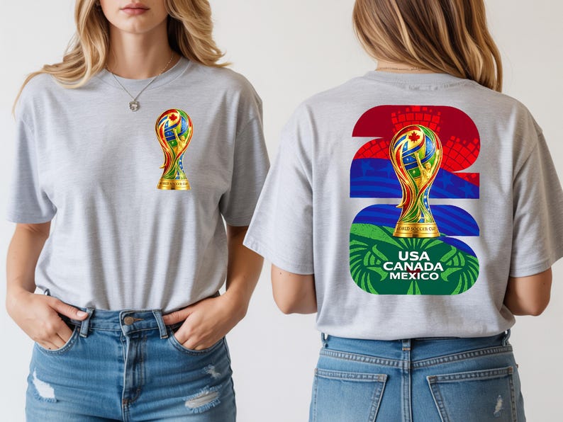 May include: A light gray t-shirt featuring a gold trophy design on the front and back. The back design includes the text "2026" and "USA CANADA MEXICO" in green, blue, and red. The trophy design incorporates various flag colors.