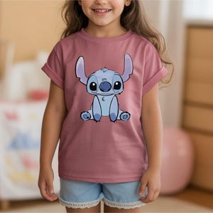Puede incluir: Una camiseta de color malva con una ilustraci&oacute;n de dibujos animados de Stitch de Lilo & Stitch. El personaje es azul con ojos y orejas grandes. La camiseta tiene las mangas enrolladas y la lleva una persona.