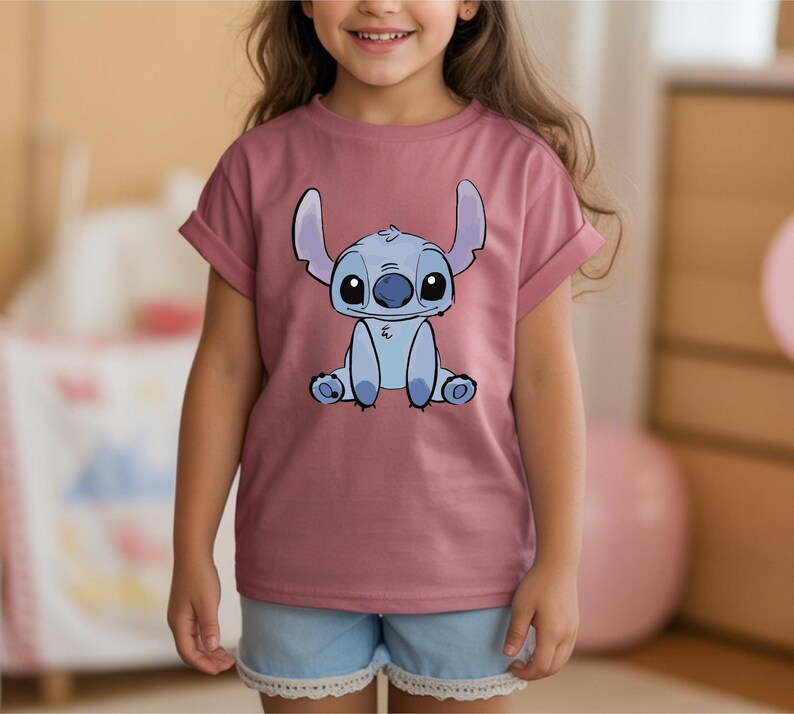 Puede incluir: Una camiseta de color malva con una ilustraci&oacute;n de dibujos animados de Stitch de Lilo & Stitch. El personaje es azul con ojos y orejas grandes. La camiseta tiene las mangas enrolladas y la lleva una persona.