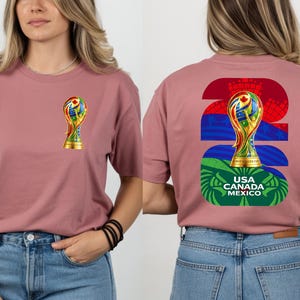 May include: Dusty rose t-shirt featuring a colorful trophy graphic on the front and back. The back design includes the number 26, a trophy, and the text "USA CANADA MEXICO". The shirt is made of a soft material and has short sleeves.