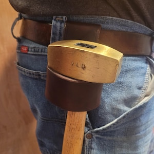 May include: A brass 2 LB (0.9 kg) mallet with a wooden handle, secured in a brown leather belt loop. The mallet is worn on the waist, with the handle pointing downwards. The mallet's head is rectangular.