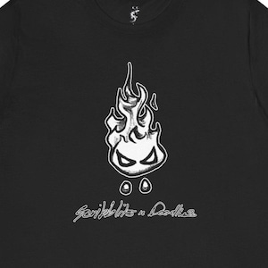 Może przedstawiać: Czarny t-shirt z białym, kresk&oacute;wkowym wzorem płomienia, z oczami i dwoma okręgami poniżej. Tekst "Scribblits & Doodles" w kursywie znajduje się pod grafiką płomienia. Koszulka wykonana jest z miękkiego materiału.