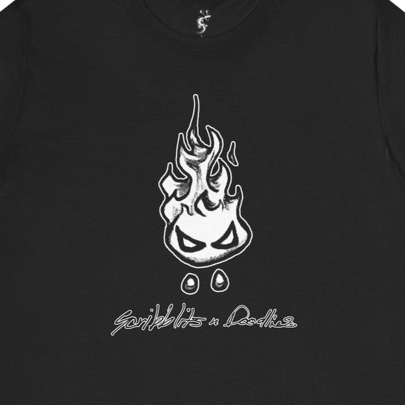 Może przedstawiać: Czarny t-shirt z białym, kresk&oacute;wkowym wzorem płomienia, z oczami i dwoma okręgami poniżej. Tekst "Scribblits & Doodles" w kursywie znajduje się pod grafiką płomienia. Koszulka wykonana jest z miękkiego materiału.