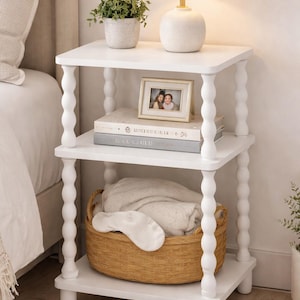 May include: A white, three-tiered side table with turned legs. The top shelf holds a small potted plant and a white lamp. The middle shelf displays books and a framed photo. A woven basket with a blanket and eye mask sits on the bottom shelf.