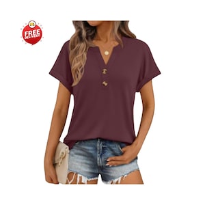 May include: Burgundy V-neck top with short sleeves and three brown buttons. The top is paired with denim shorts. A gold necklace is visible. A small, off-white clutch is held in the hand.