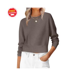 Op de afbeelding: Een bruine trui met ronde hals, lange mouwen en een geribbelde zoom. De trui wordt gecombineerd met een witte broek. Een ketting met een hanger is zichtbaar. De afbeelding bevat een "GRATIS VERZENDING"-logo.