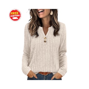 May include: A cream-colored, long-sleeved top with a ribbed texture and a V-neckline. The top features two brown buttons and is paired with blue jeans and a brown belt. The image also includes a "FREE SHIPPING" logo.