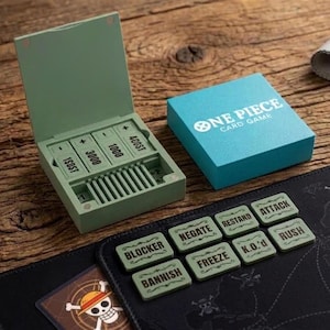 Op de afbeelding: Een saliegroene plastic speldoos met open deksel, met numerieke fiches gelabeld 1000, 3000 en 4000. Daarnaast staat een turquoise doos met de tekst ONE PIECE CARD GAME en acht saliegroene rechthoekige fiches met de tekst BLOCKER, NEGATE, RESTAND, ATTACK, BANNISH, FREEZE, K.O.'d en RUSH.