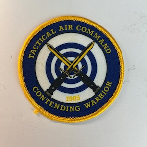 May include: A circular embroidered patch with a yellow border, a blue outer ring, and a white center featuring a blue target design. Two crossed black and yellow daggers are centered. Text reads TACTICAL AIR COMMAND CONTENDING WARRIOR 1985.