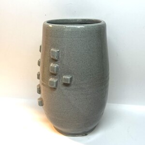 Puede incluir: Un jarr&oacute;n de cer&aacute;mica gris con una superficie texturizada y una serie de acentos cuadrados en relieve. El jarr&oacute;n tiene una forma cil&iacute;ndrica con una parte superior ligeramente ensanchada. El color es gris apagado.