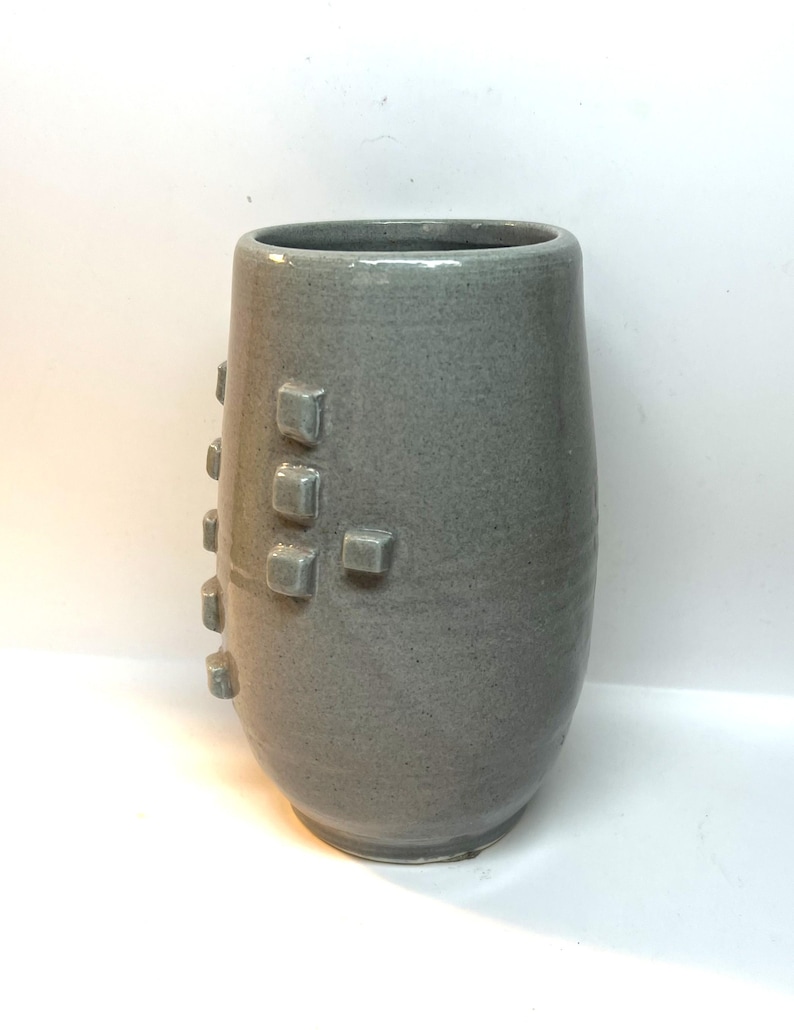Puede incluir: Un jarr&oacute;n de cer&aacute;mica gris con una superficie texturizada y una serie de acentos cuadrados en relieve. El jarr&oacute;n tiene una forma cil&iacute;ndrica con una parte superior ligeramente ensanchada. El color es gris apagado.