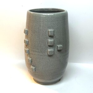 May include: A gray ceramic vase with a textured surface and a series of square, raised accents. The vase has a cylindrical shape with a slightly wider base and a smooth, open top. The color is a muted gray.