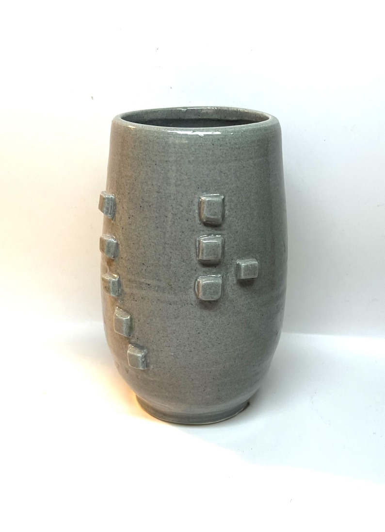 Puede incluir: Un jarr&oacute;n de cer&aacute;mica gris con una superficie texturizada y una serie de acentos cuadrados en relieve. El jarr&oacute;n tiene una forma cil&iacute;ndrica con una base ligeramente m&aacute;s ancha y una parte superior lisa y abierta. El color es gris apagado.