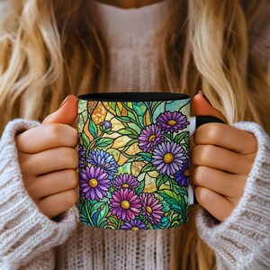 May include: A black coffee mug featuring a stained-glass floral design with purple, blue, and yellow flowers. The mug has a black interior and is held by a person wearing a white sweater.