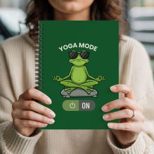 Peut inclure: Un cahier vert à spirales avec les mots "YOGA MODE" au-dessus d'une grenouille de dessin animé portant des lunettes de soleil et méditant. En dessous, un bouton "ON". Le cahier est tenu par une personne.