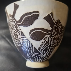 May include: A ceramic bowl with a cream-colored base featuring a dark brown, hand-carved graphic pattern of a giraffe. The design incorporates intricate, flowing lines that define the animal's spots and facial features against the light background.