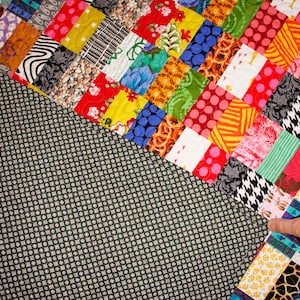 May include: A colorful patchwork quilt with a variety of fabrics and patterns. The quilt is made up of squares and rectangles of different colors and designs. The quilt is sewn together with a dark green backing.