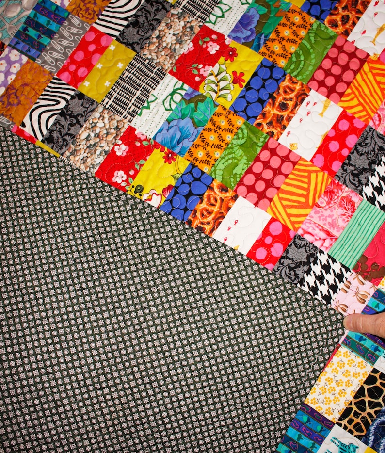 May include: A colorful patchwork quilt with a variety of fabrics and patterns. The quilt is made up of squares and rectangles of different colors and designs. The quilt is sewn together with a dark green backing.