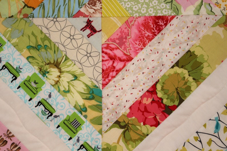 May include: Close-up of a colorful quilt with a diagonal pattern. The quilt features various fabric patterns, including floral, geometric, and cat motifs. The color palette includes green, pink, blue, and yellow.