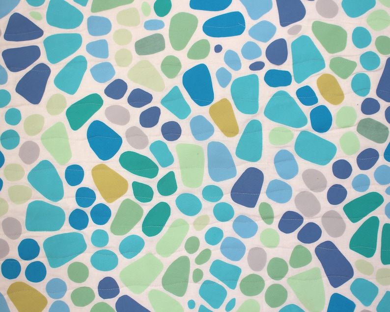 May include: A close-up of a fabric with a pattern of irregular shapes in shades of blue, green, and yellow on a white background. The shapes vary in size and are scattered across the surface. The fabric has a quilted appearance.
