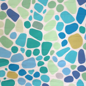 May include: A close-up of a fabric with a pattern of irregular shapes in shades of blue, green, and yellow on a white background. The shapes vary in size and are outlined with a stitched pattern. The overall design is reminiscent of pebbles or abstract organic forms.