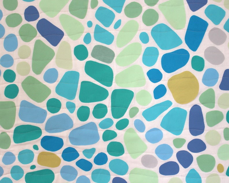 May include: A close-up of a fabric with a pattern of irregular shapes in shades of blue, green, and yellow on a white background. The shapes vary in size and are outlined with a stitched pattern. The overall design is reminiscent of pebbles or abstract organic forms.